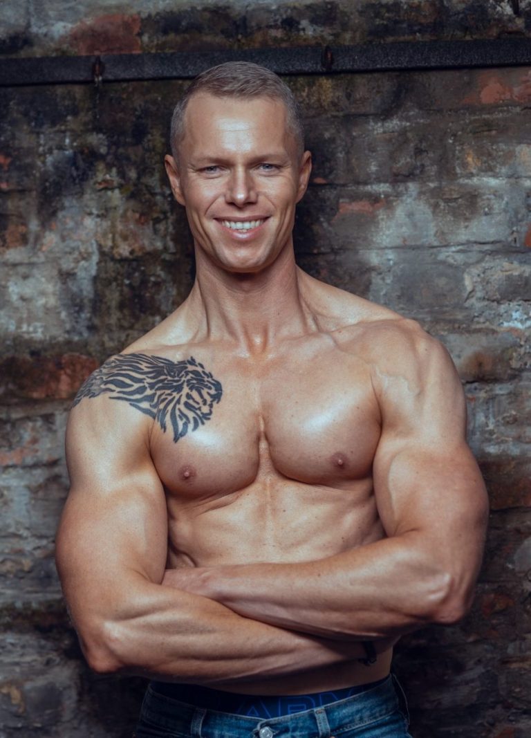 Mann mit muskulösem Körper und Tiger-Tattoo auf dem Oberarm, lächelt vor Backsteinwand.